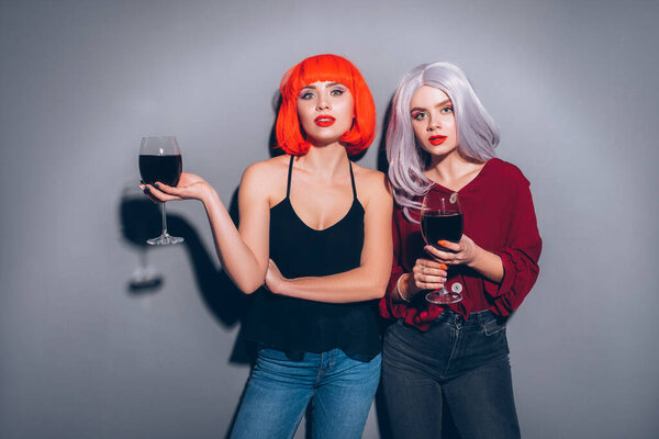 Две милые пьяные девушки в красных и серых париках, одетые в джинсы и блузки на нейтральном сером фоне. Они с радостью пьют вино. Улыбающийся портрет с резким освещением моды