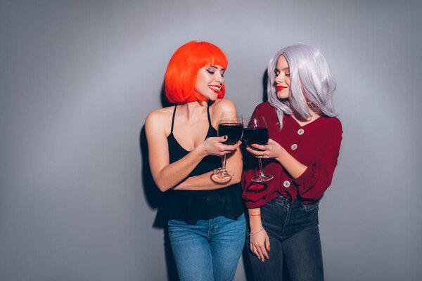 Две милые пьяные девушки в красных и серых париках, одетые в джинсы и блузки на нейтральном сером фоне. Они с радостью пьют вино. Улыбающийся портрет с резким освещением моды