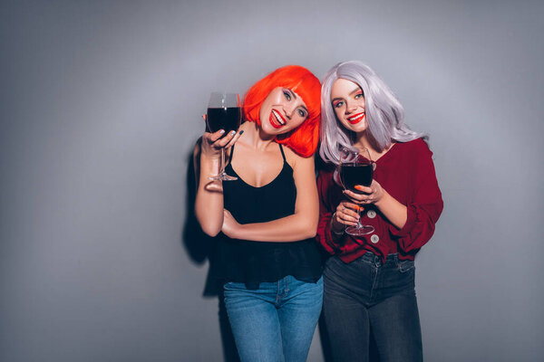 Две милые пьяные девушки в красных и серых париках, одетые в джинсы и блузки на нейтральном сером фоне. Они с радостью пьют вино. Улыбающийся портрет с резким освещением моды