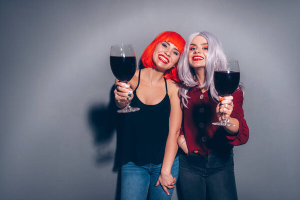 Две милые пьяные девушки в красных и серых париках, одетые в джинсы и блузки на нейтральном сером фоне. Они с радостью пьют вино. Улыбающийся портрет с резким освещением моды