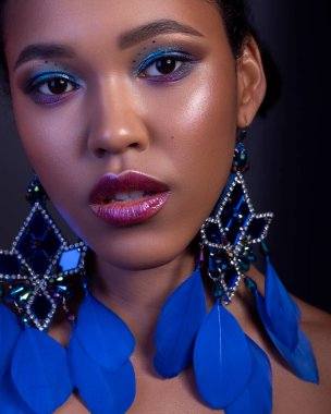 Koyu tenli, genç ve güzel bir kadın koyu bir arka planda şehvetle poz veriyor. Parlak renkli, yaratıcı makyajlı kusursuz bir cilt. Kocaman egzotik küpeler takıyor..