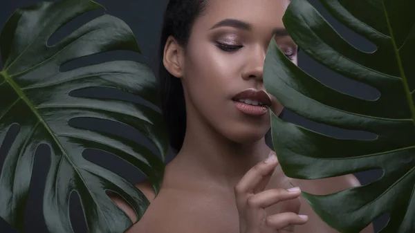 Dark skinned beautiful young woman posing sensually on a neutral green background with monstera leafs. Flawless skin with nude make up. Skin care concept portait with space for text.