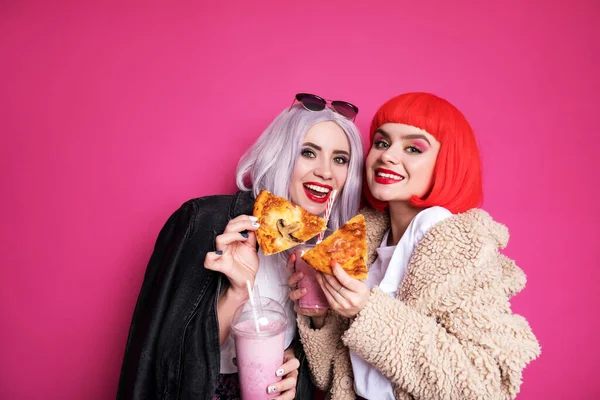 ピンクの背景に立つ10代の女の子のような2人のかわいい女の子の友人の女性 カラフルなウィッグとスタイリッシュなカジュアルな衣装を着て ミルクセーキでピザを食べています 幸せな感情的な肖像画 — ストック写真