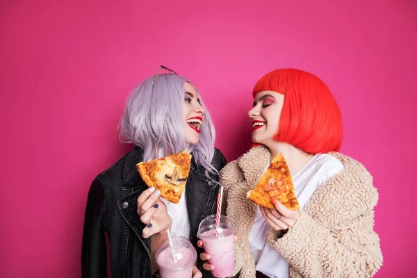 ピンクの背景に立つ10代の女の子のような2人のかわいい女の子の友人の女性 カラフルなウィッグとスタイリッシュなカジュアルな衣装を着て ミルクセーキでピザを食べています 幸せな感情的な肖像画 — ストック写真