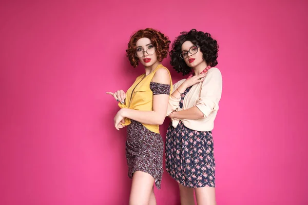 ピンクの背景に立ってオタク会計士のように見える2人のかわいい女の子の友人の女性 彼らは巻きブルネットのウィッグとスタイリッシュなレトロカジュアル衣装を身に着けている — ストック写真