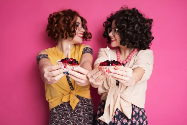 两个可爱的女友站在粉红的背景上 看起来像个书呆子会计 他们穿着卷曲的黑发假发和过时的复古休闲装 手中拿着蜡烛的浆果蛋糕 — 图库照片