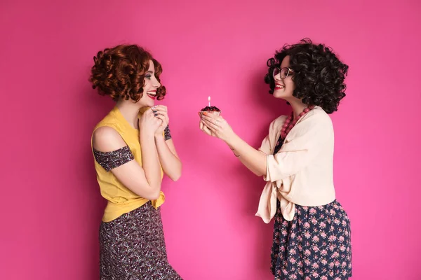ピンクの背景に立ってオタク会計士のように見える2人のかわいい女の子の友人の女性 彼らは巻きブルネットのウィッグとスタイリッシュなレトロカジュアルな衣装を着ています 手にキャンドル付きベリーカップケーキ — ストック写真