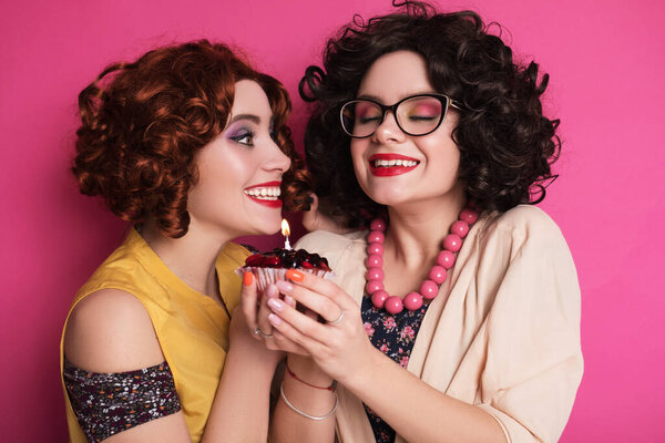 Two cute girl friends woman looking like nerd accountants standing on a pink background. They wear curly brunette wigs and unstylish retro casual outfits. Berry cupcake with candle in their hands.