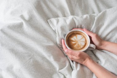 Kadın elleri, beyaz bardakta kahve tutarken gri çarşaf ekiyor. Latte sanatı en üstte. Metin için boşluk