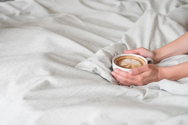 Kadın elleri, beyaz bardakta kahve tutarken gri çarşaf ekiyor. Latte sanatı en üstte. Metin için boşluk