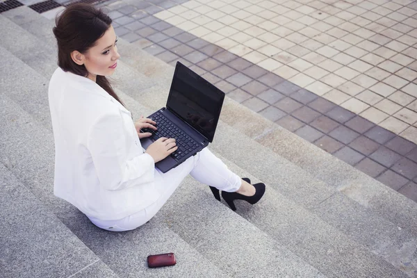 Belle femme d'affaires brune en costume blanc avec ordinateur portable sur ses genoux, dactylographiant, travaillant à l'extérieur. Espace de copie — Photo