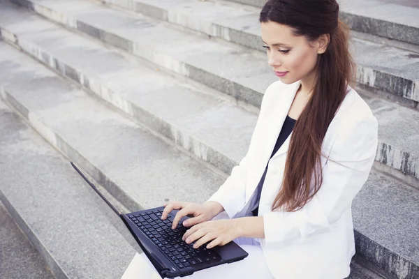 Belle femme d'affaires brune en costume blanc avec ordinateur portable sur ses genoux, dactylographiant, travaillant à l'extérieur. Espace de copie — Photo