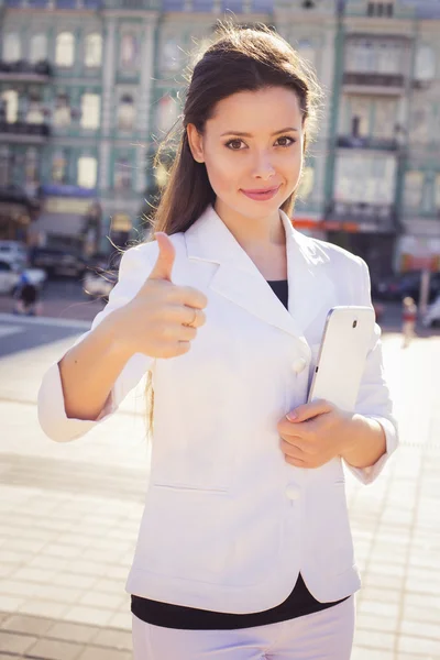 Belle femme d'affaires brune en costume blanc avec tablette dans ses mains à l'extérieur montrant pouce vers le haut — Photo