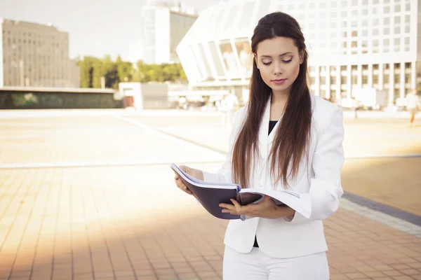 Beautiful brunette business woman in white suit with folder of documents in her hands reading outdoors. copy space — Stock Photo, Image