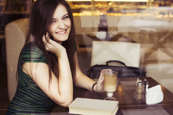 Mujer morena sentada en el café leyendo libro, tachonado y tomando café y hablando por teléfono. Copiar espacio — Foto de Stock