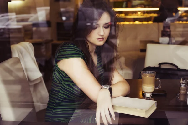 Mujer morena sentada en el café leyendo libro, tachonado y tomando café y hablando por teléfono. Copiar espacio — Foto de Stock