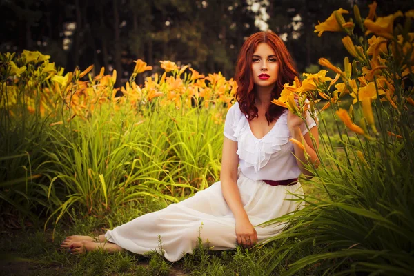 Beautiful brunette caucasian woman in white dress at the park in red and yellow flowers on a summer sunset holding flowers sitting on the grass. Copy Space — Stock Photo, Image