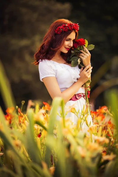 Beautiful brunette caucasian woman in white dress at the park in red and yellow flowers on a summer sunset holding roses — Stock Photo, Image