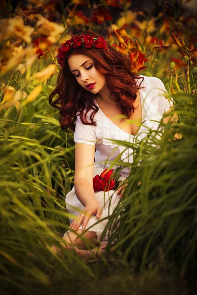 Beautiful brunette caucasian woman in white dress at the park in red and yellow flowers on a summer sunset holding roses — Stock Photo, Image