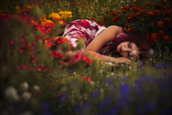 Beautiful brunette caucasian woman in dress at the park in flowers on a summer sunset holding flowers laying on the grass — Stock Photo, Image