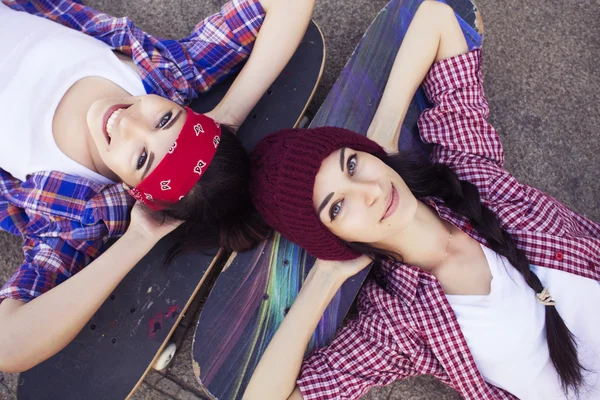 Due amiche adolescenti brune in costume da hipster (pantaloncini jeans, keds, camicia a quadri, cappello) con uno skateboard al parco all'aperto . — Foto Stock