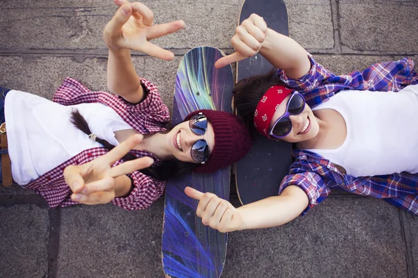 Two Brunette teenage girls friends in hipster outfit (jeans shorts, keds, plaid shirt, hat) with a skateboard at the park outdoors. — Stock Photo, Image