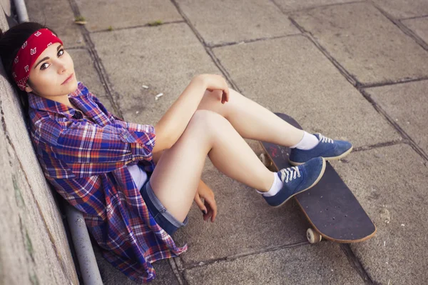 Gadis berambut coklat dengan pakaian hipster (celana jeans pendek, keds, kemeja kotak-kotak, topi) dengan skateboard di taman luar ruangan. Salin ruang — Stok Foto