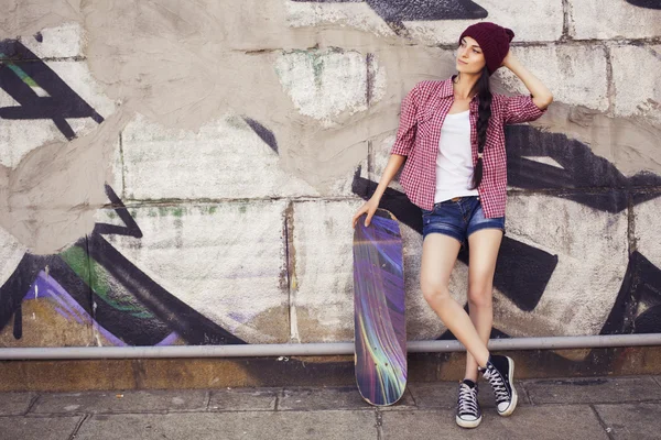 Morena adolescente en traje hipster (pantalones cortos vaqueros, camas, camisa a cuadros, sombrero) con un monopatín en el parque al aire libre. Copiar espacio — Foto de Stock