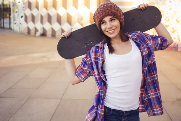 Brunette tiener meisje in hipster outfit (jeans broek, keds, plaid shirt, hoed) met een skateboard het park in de open lucht. kopie ruimte — Stockfoto