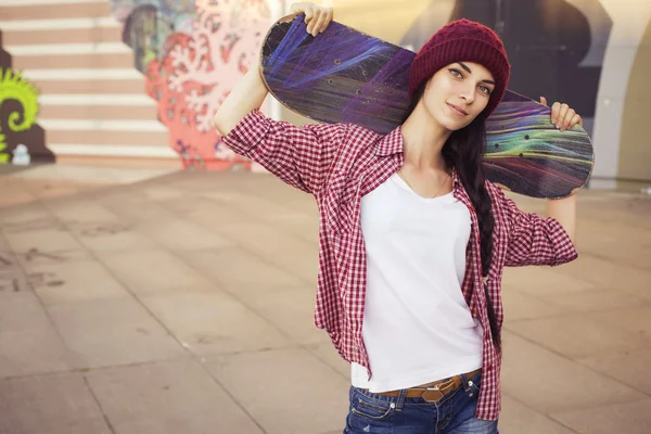 Bruna adolescente in abito hipster (pantaloncini jeans, keds, camicia a quadri, cappello) con uno skateboard al parco all'aperto. Copia spazio — Foto Stock