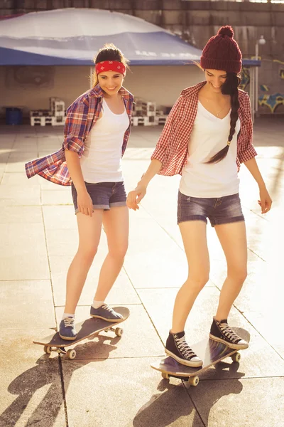 Duas meninas adolescentes morena amigos em roupa hipster (calções de ganga, keds, camisa xadrez, chapéu) com um skate no parque ao ar livre . — Fotografia de Stock