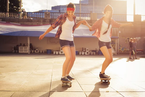 Due amiche adolescenti brune in costume da hipster (pantaloncini jeans, keds, camicia a quadri, cappello) con uno skateboard al parco all'aperto. Copia spazio — Foto Stock