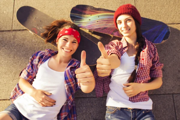 Två brunett tonårsflickor vänner i hipster outfit (jeansshorts, keds, rutiga skjortan, hatt) med en skateboard på park utomhus. — Stockfoto