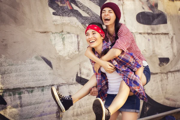 Due amiche adolescenti brune in costume da hipster (pantaloncini jeans, keds, camicia a quadri, cappello) con uno skateboard al parco all'aperto. Copia spazio — Foto Stock