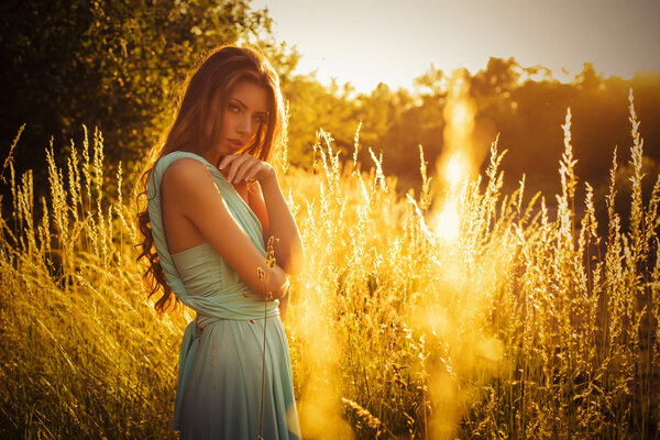 Beautiful blonde with a long curly hair in a long evening dress in motion outdoors in nature in summer sunset. Copy Space