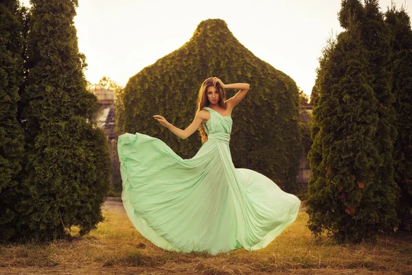 Beautiful blonde with a long curly hair in a long evening dress in motion outdoors near retro vintage building in summer sunset. Copy Space — Stock Photo, Image