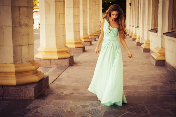 Hermosa rubia con un largo pelo rizado en un largo vestido de noche en movimiento al aire libre cerca del edificio retro vintage en la puesta de sol de verano. Copiar espacio — Foto de Stock