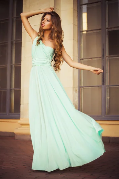 Hermosa rubia con un largo pelo rizado en un largo vestido de noche en movimiento al aire libre cerca de edificio retro vintage todo en hojas en la puesta de sol de verano — Foto de Stock