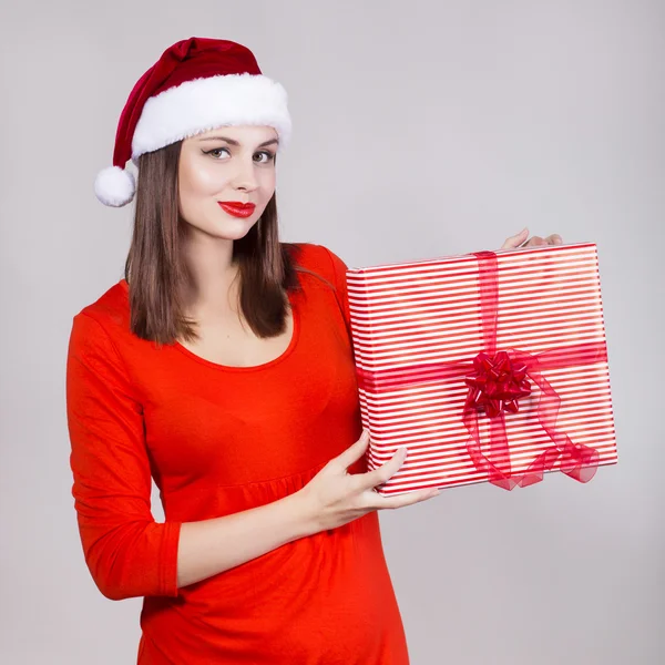 Bruneta žena v červené šaty a klobouk Santa drží současnost a hledět v kameře — Stock fotografie