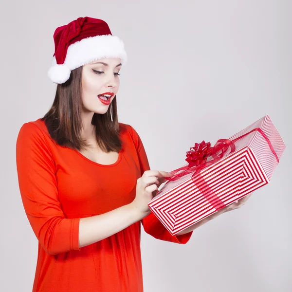 Bruneta žena v červené šaty a klobouk Santa drží současnost a hledět v kameře — Stock fotografie