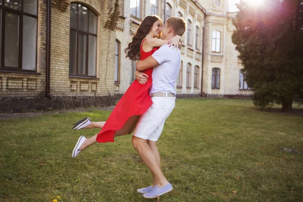 Piękna brunetka para zakochanych przytulanie na randkę w parku. Ona w czerwonej sukience, zwinięte fryzurę i makijaż. On w koszulę i spodnie. Stylu casual. Na zewnątrz. Ciepła Pogoda, zachód słońca — Zdjęcie stockowe