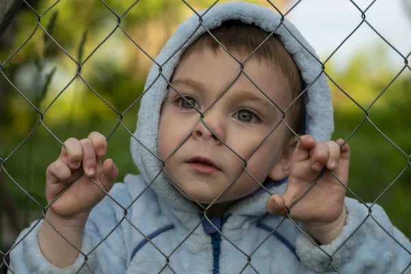 The child stands holding on to the mesh fence leaning against him face