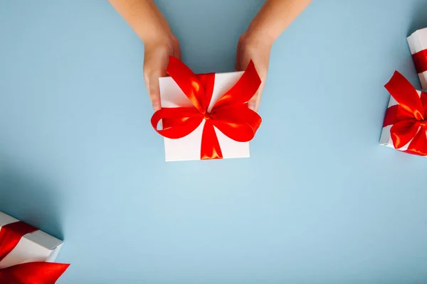Le mani dei bambini tengono una scatola regalo bianca con un fiocco rosso su uno sfondo blu. — Foto Stock