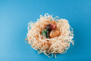 Üç paskalya yumurtası kırmızı, sarı ve yeşile boyanır. Yumurtalar odun talaşı yuvasında yatar. Mavi arka planda boyanmış paskalya yumurtaları.. 