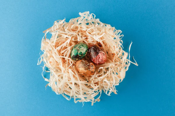 Üç paskalya yumurtası kırmızı, sarı ve yeşile boyanır. Yumurtalar odun talaşı yuvasında yatar. Mavi arka planda boyanmış paskalya yumurtaları. Düz yatıyordu. Boşluğu kopyala