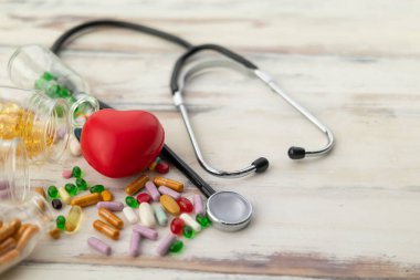 Kırmızı kalbin üst görüntüsü ilaç, vitamin ve kaymak gibi ahşap zeminde steteskop ile çevrili.