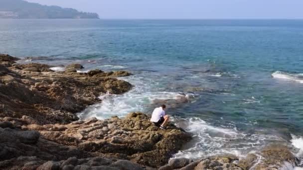 Un joven está de pie en una costa rocosa y filmando las olas golpeando las rocas — Vídeo de stock
