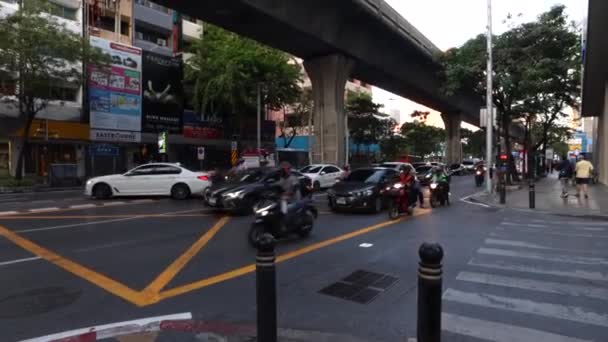 Ocupado camino a lo largo del cual los coches y scooters conducen en Bangkok. La gente camina por las aceras y el metro cruza el puente de la carretera — Vídeo de stock
