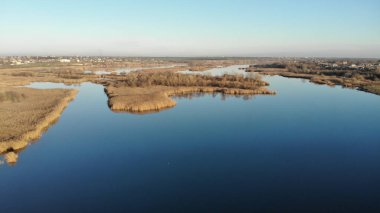 Mavi suyu olan bir nehrin haliçleri. Nehrin kıyısında ve ortasında kuru otlar ve sazlıklar var. Küçük evleri olan bir köy var. 