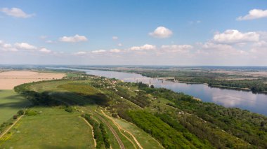 Ağaçlı yeşil tarlalar ve orman ve demiryolu köprüsünün geçtiği bir nehir.